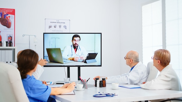 Medical team holding online conference in boardroom with man specialist doctor and taking notes on clipboard. Group of doctors discussing diagnosis about treatment of patients using video call