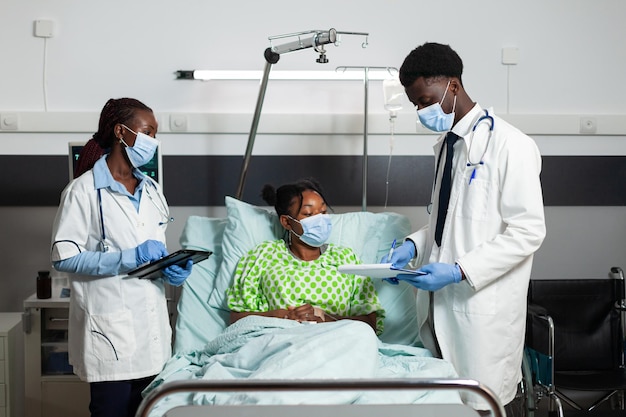 Medical team with protective face masks to prevent infection with covid19 monitoring sick patient during clinical appointment in hospital ward. African american doctors explaining healthcare treatment