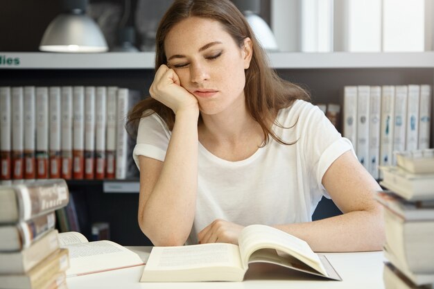 Medical University Caucasian female student studying at library, beautiful college woman sleeping while sitting in front of an open book resting her chin on a hand, looking exhausted.