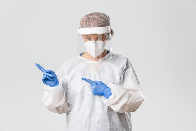 Медицинские работники, пандемия covid-19, концепция коронавируса. Удивленная женщина-врач, ученый или техническая лаборатория в средствах индивидуальной защиты выглядят изумленными, указывая пальцем в верхнем левом углу