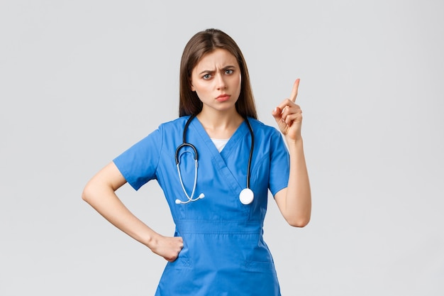 医療従事者、ヘルスケア、covid-19および予防接種の概念。青いスクラブで若い女性の看護師や医師をしかめっ面、眉をひそめている、患者の悪い行動を叱るような指を振る、灰色の背景