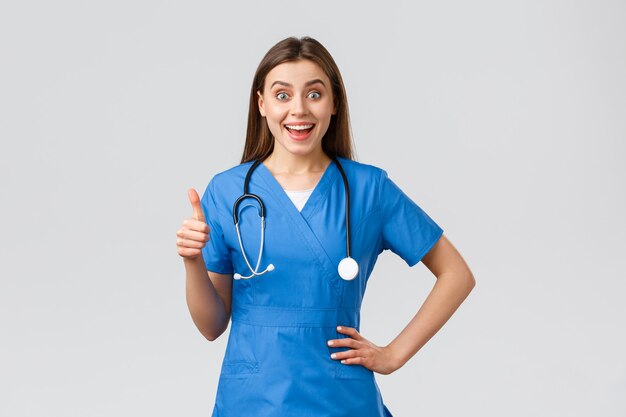 Медицинские работники, здравоохранение, covid-19 и концепция вакцинации. Удивленная взволнованная медсестра или врач в синих скрабах со стетоскопом, одобрительно показывает большие пальцы вверх, как идея, оптимистично улыбается