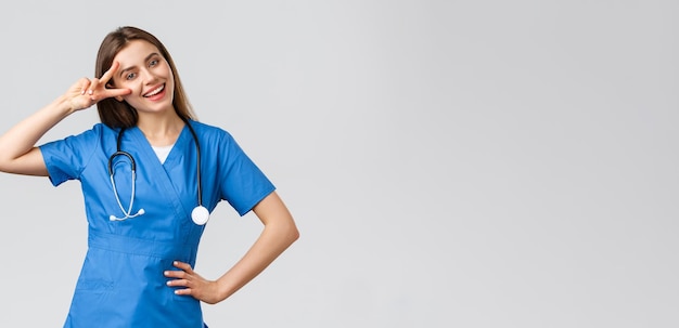 Medical workers healthcare covid19 and vaccination concept Cheerful attractive female nurse or doctor in blue scrubs stethoscope show peace sign stay optimistic standing grey background