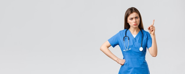 Medical workers healthcare covid19 and vaccination concept Frowning young female nurse or doctor in blue scrubs frowning shaking finger as scolding patient bad behaviour grey background