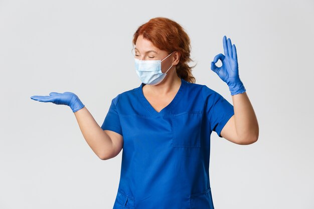 Medical workers,  pandemic, coronavirus concept. Happy smiling female doctor, vet or physician in face mask and gloves, holding something on palm and show okay in approval, recommend