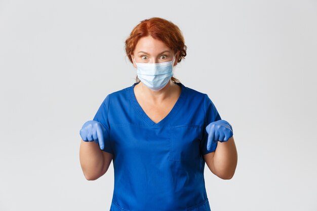 의료 노동자, 전염병, 코로나 바이러스 개념. 충격적이고 흥분된 빨간 머리 중년 여성 의사, 의사가 손가락을 아래로 가리키고 큰 뉴스를 전하고 놀란 표정, 회색 벽