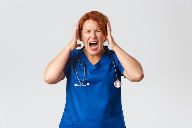 Medicine, healthcare and coronavirus concept. Annoyed and angry, screaming female doctor, nurse going nuts, shouting and shaking hands, losing temper and feeling mad, standing over grey wall.