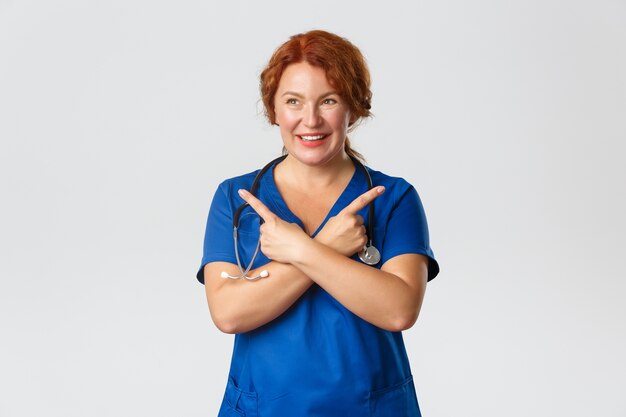 Medicine, healthcare and coronavirus concept. Cheerful smiling middle-aged redhead doctor, physician or nurse in blue scrubs showing way, medical worker pointing sideways, choosing.