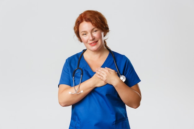 Medicine, healthcare and coronavirus concept. Touched and sympathetic redhead nurse, middle-aged doctor or physician in scrubs, press hands to heart and smiling flattered, being praised