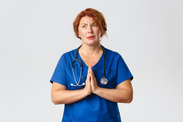 Medicine, healthcare and coronavirus concept. Worried and hopeful redhead female medical worker hoping for end of  pandemic, praying or pleading with hands clasped together.