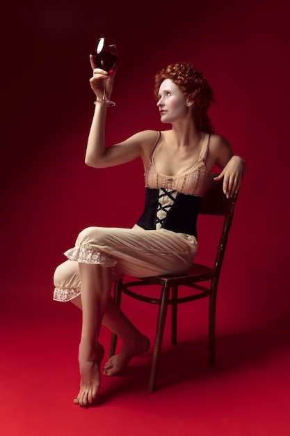 Medieval redhead young woman as a duchess in black corset and night clothes sitting on a chair on red space with a glass of wine