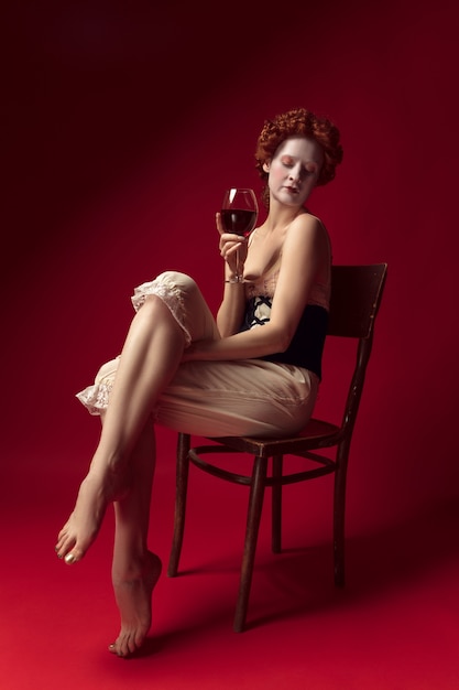 Medieval redhead young woman as a duchess in black corset and night clothes sitting on a chair on red space with a glass of wine