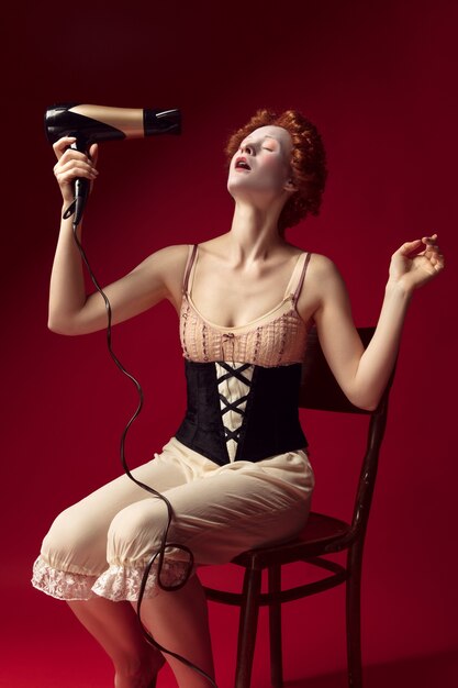 Medieval redhead young woman as a duchess in black corset and night clothes sitting on the chair on red wall. Doing her hair with dryer. Concept of comparison of eras, modernity and renaissance.