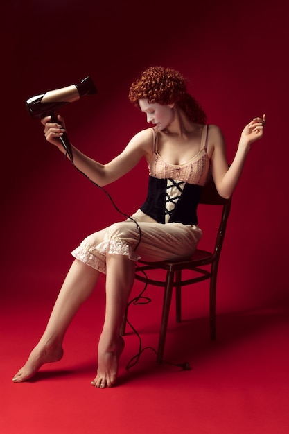 Free photo medieval redhead young woman as a duchess in black corset and night clothes sitting on the chair on red wall. doing her hair with dryer. concept of comparison of eras, modernity and renaissance.