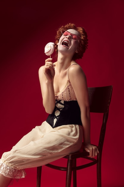 Medieval redhead young woman as a duchess in black corset, sunglasses and night clothes sitting on a chair on red space with a candy