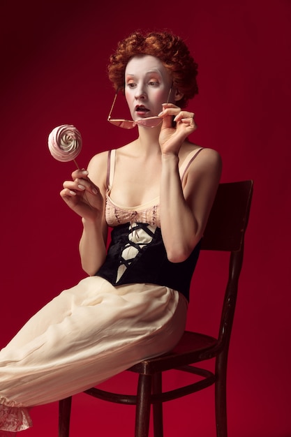 Medieval redhead young woman as a duchess in black corset, sunglasses and night clothes sitting on a chair on red wall with a candy. concept of comparison of eras, modernity and renaissance.