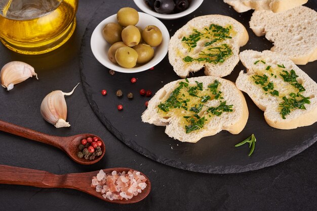 Mediterranean snacks set. Olives, oil, herbs and sliced ciabatta bread on black slate stone board over painted dark blue surface, top view. Flat lay.