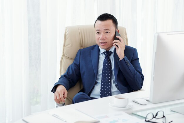 彼のオフィスの机に座って電話をかけるアジア系のビジネスマンの中程度のクローズアップ