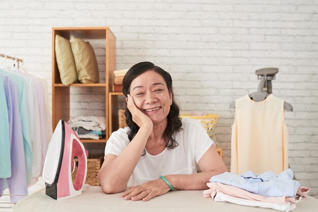 Medium closeup of Asian senior wowan enjoying housework leaning on ironboard and smiling cheerfully