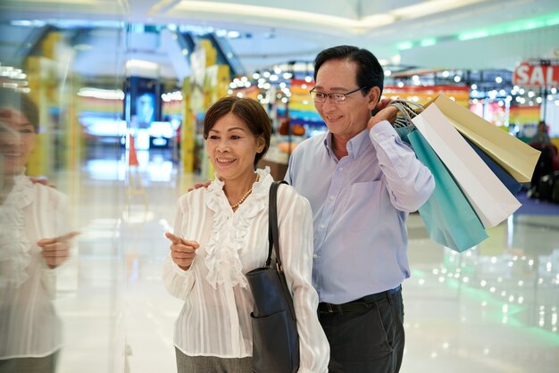 Medium shot of Asian middle aged couple window shopping at a mall, man holding  shop bags