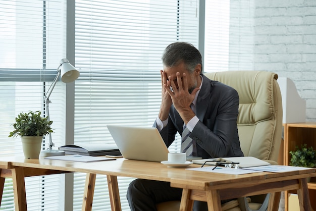 Medium shot of businessman seated at desk with his hands on his face frustrated by failure