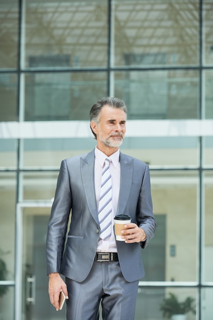 Medium shot of smart business executive in forlmalwear standing outside the office building to have his takeaway coffee