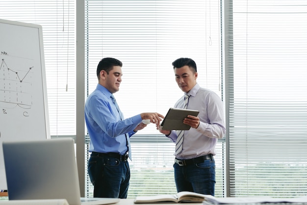 Medium shot of two colleagues standing in the office and discussing data on tablet PC