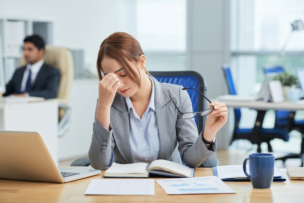 Medium shot of young Asian woman sitting at desk in office and rubbing nose