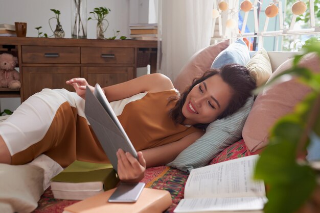 Medium shot of young woman resting in bed reading the ebook