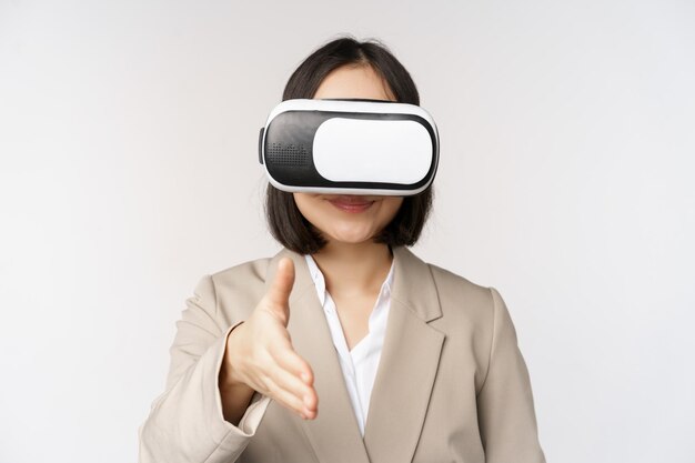 Meeting in vr chat Asian businesswoman in virtual reality glasses extending hand for handhshake with business partner greeting someone standing over white background