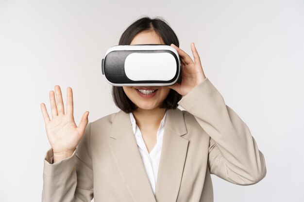 Meeting in vr chat Asian businesswoman in virtual reality glasses raising hand and saying hello greeting someone standing over white background