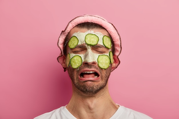 Melancholy male model has homemade mask with cucumbers on face, tired of visiting spa salon, wears white t shirt and bath hat, cannot feel relaxed