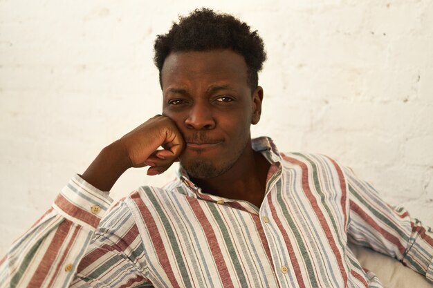 Melancholy, sadness and depression concept. Close up shot of sad upset young dark skinned guy looking at camera with thinned lips having sorrowful facial expression