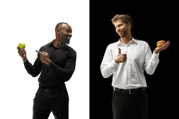 Men eating a hamburger and fresh fruits on a black and white background. the happy afro and caucasian men. the burger, fast, healthy and unhealthy food concept