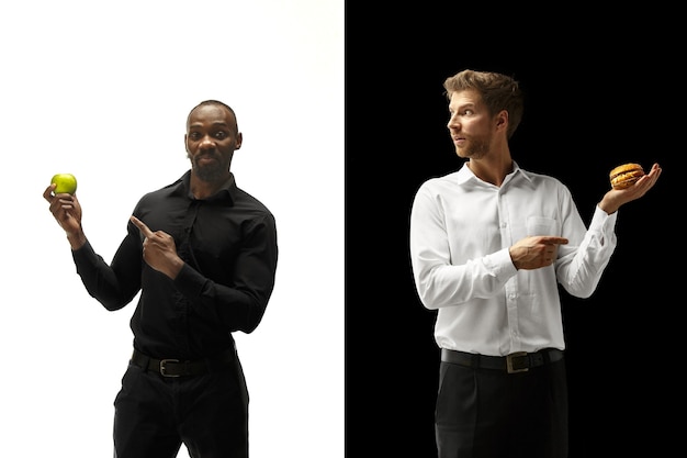 Men eating a hamburger and fresh fruits on a black and white background. The happy afro and caucasian men. The burger, fast, healthy and unhealthy food concept