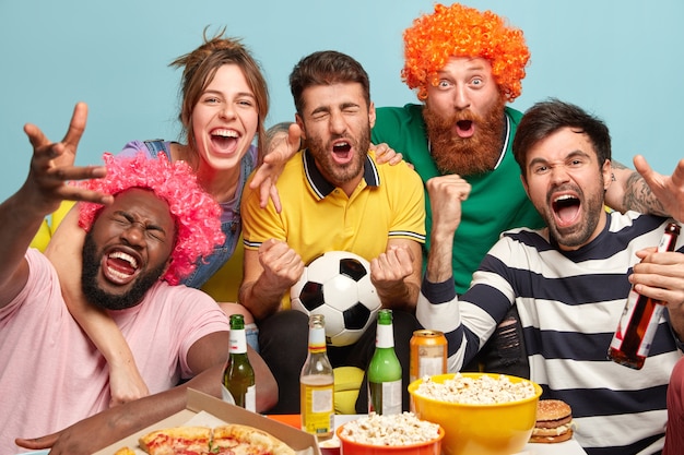 Men and woman fans watch football on TV at home, enjoy exciting game, clench fists, celebrate victory, express positive emotions, have popcorn in bowls, eat pizza, pose over blue wall.