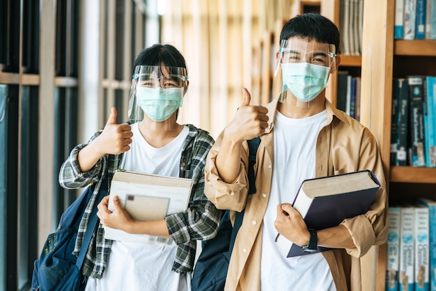 Free photo men and women wear masks to stand, hold books in the library and put their thumbs up.