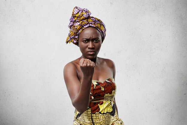 Menacing black woman with big eyes and full lips wearing colorful scarf on head and dress showing her fist looking angry and furious threatening with gesture. Emotional dark-skinned woman