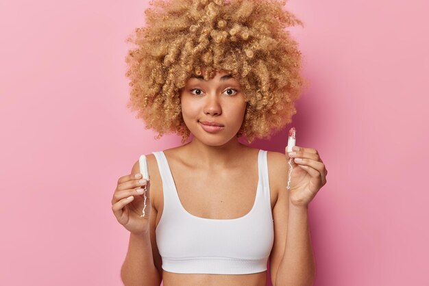 Menstrual cycle concept Young hesitant woman with curly hair holds two tampons for periods dressed in white cropped top isolated over pink background cares about intimate hygiene has bleeding