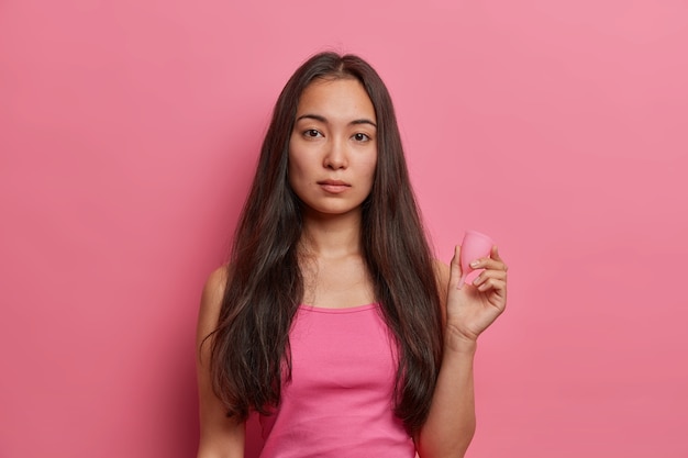 Menstrual protection, gynecology and feminine hygiene concept. Serious brunette ethnic woman holds menstrual cup, uses zero waste absorbent product in blood period, isolated on pink wall