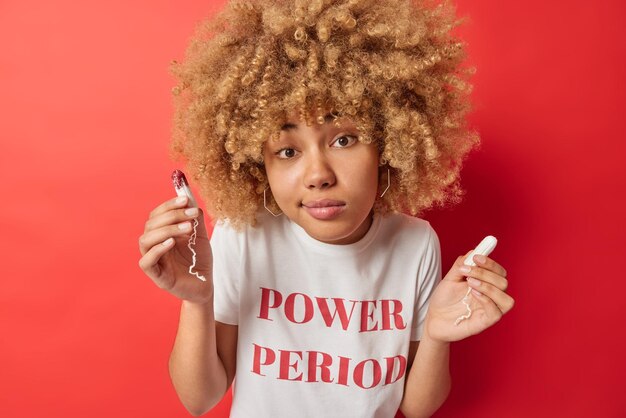 Menstruation gynecology and hygiene concept Young curly haired European woman holds two tampons chooses best absorbency product wears white t shirt with inscription isolated over red background