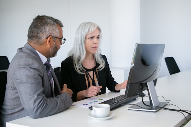 Mentor helping intern at workplace. Colleagues watching content on pc monitor, sitting at table with paper diagram. Business communication concept