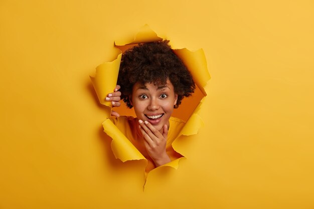 Merry brunette adult woman holds chin, smiles broadly, shows good dental treatment, has healthy skin, looks curiously happily through ripped paper hole, yellow bright background