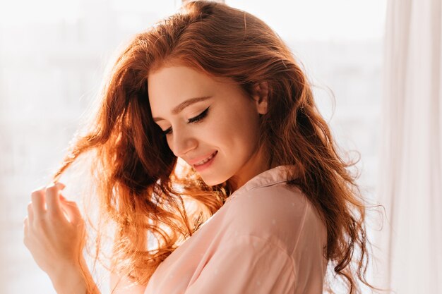 Merry caucasian girl playing with her wavy hair. Indoor photo of stunning lady enjoying photoshoot at home.