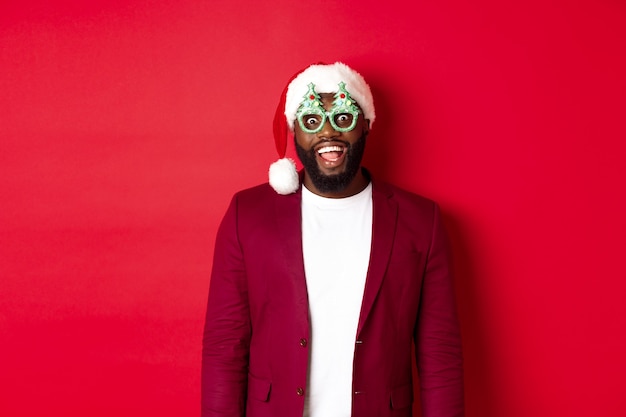 Free photo merry christmas. cheerful black man wearing funny party glasses and santa hat, smiling joyful, celebrating winter holidays, standing over red background.