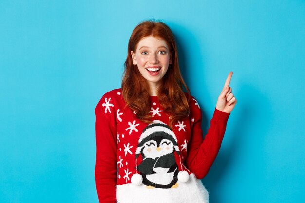 Merry Christmas. Cheerful redhead girl in xmas sweater, pointing finger at upper right corner and smiling excited, showing new year promo.