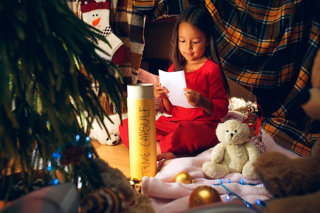 Buon natale e buone feste. la bambina sveglia del bambino scrive la lettera a babbo natale vicino all'albero di natale