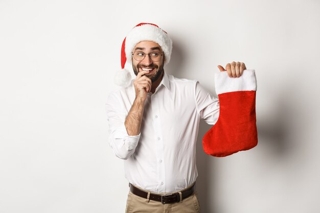 Merry christmas, holidays concept. Adult man looking happy and curious at xmas sock, receive gifts, wearing santa hat 
