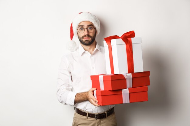 Merry christmas, holidays concept. Confused man in Santa hat holding pile of presents, found gifts under xmas tree, standing against white background.