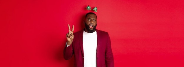 Free photo merry christmas masculine black man in costume and silly party headband showing peace sign but looki